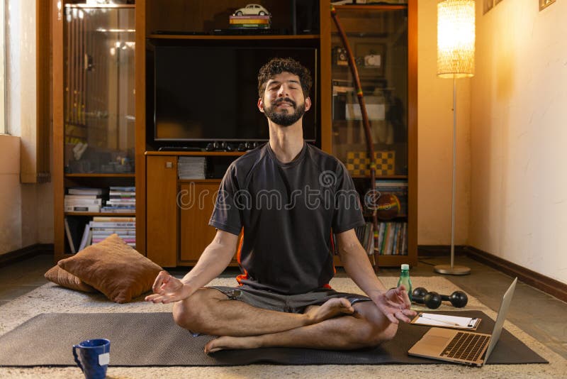 Young man performing meditation, listening to lessons through his computer, sitting in lotus pose at his home. Yoga concept. Young man performing meditation, listening to lessons through his computer, sitting in lotus pose at his home. Yoga concept.