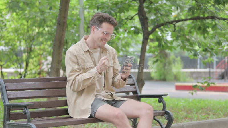 Jovem moderno celebrando no smartphone enquanto sentado no banco