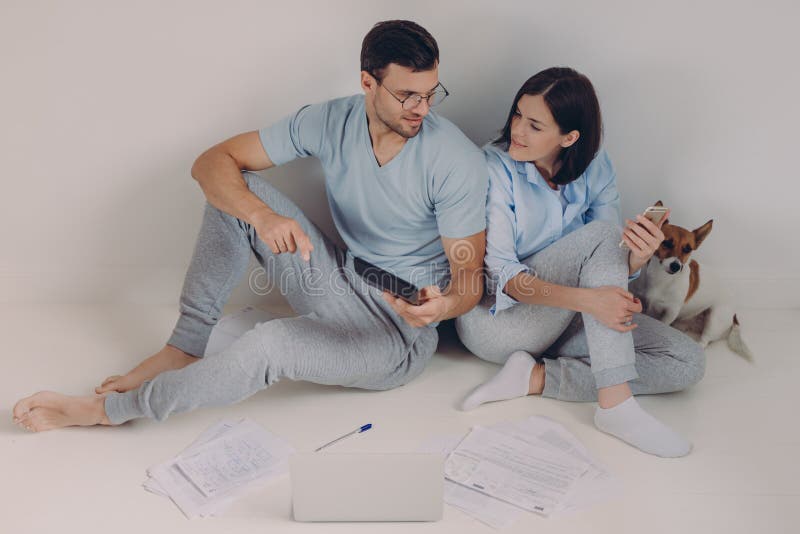A jovem mulher alegre usa depositar o app em seu telefone celular, seu marido mostra figuras na calculadora, cercada com papéis, imagens de stock royalty free A jovem mulher alegre usa depositar o app em seu telefone celular, seu marido mostra figuras na calculadora, cercada com papéis, imagens de stock royalty free