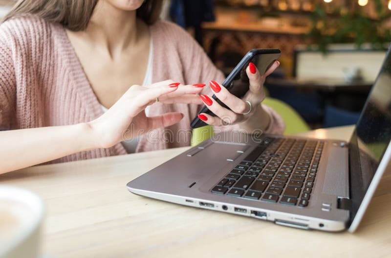A jovem mulher atrativa senta-se em uma tabela em um caf? com uma x?cara de caf? e um port?til e usa-se o telefone fotos de stock A jovem mulher atrativa senta-se em uma tabela em um caf? com uma x?cara de caf? e um port?til e usa-se o telefone fotos de stock
