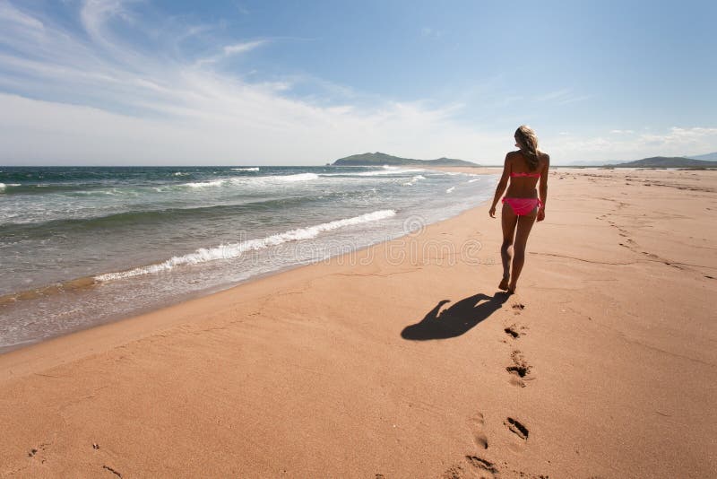 A jovem mulher atravessa à distância a praia vazia, selvagem contra um céu azul, a areia amarela e o mar Grande ângulo fotografia de stock royalty free A jovem mulher atravessa à distância a praia vazia, selvagem contra um céu azul, a areia amarela e o mar Grande ângulo fotografia de stock royalty free