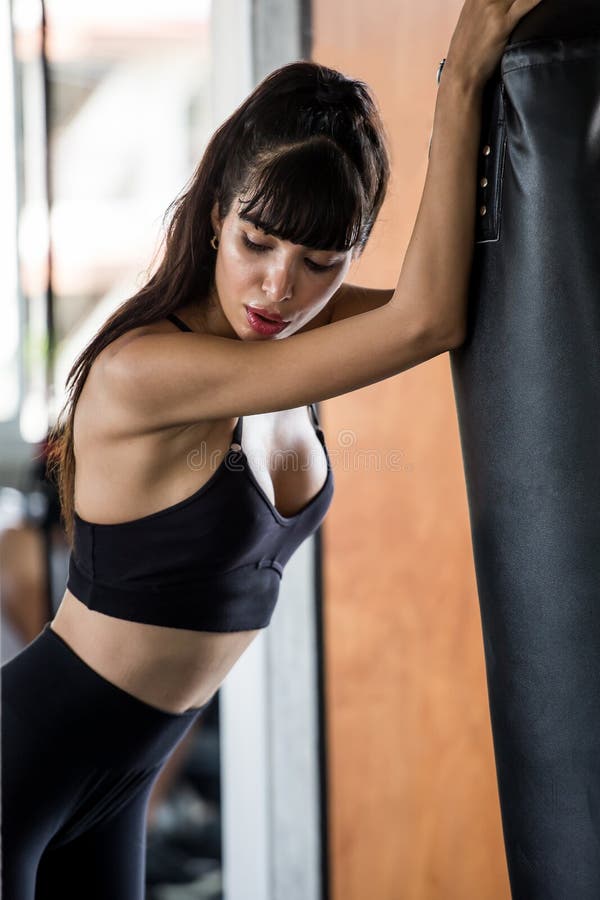 Jovem mulher bonita do pugilista cansado tomando uma ruptura do exercício que inclina-se contra o saco de perfuração no gym da ap fotos de stock Jovem mulher bonita do pugilista cansado tomando uma ruptura do exercício que inclina-se contra o saco de perfuração no gym da ap fotos de stock