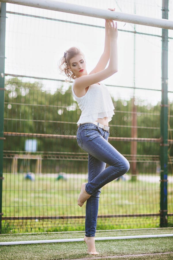 Jovem mulher bonita em um campo de futebol verde Menina que está na porta do futebol, vestida na calças de ganga, um t-shirt bran imagem de stock Jovem mulher bonita em um campo de futebol verde Menina que está na porta do futebol, vestida na calças de ganga, um t-shirt bran imagem de stock