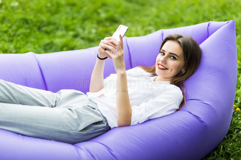 A jovem mulher bonita que encontra-se no uso inflável app do lamzac do sofá do telefone, consulta o Internet ao descansar na gram fotografia de stock A jovem mulher bonita que encontra-se no uso inflável app do lamzac do sofá do telefone, consulta o Internet ao descansar na gram fotografia de stock