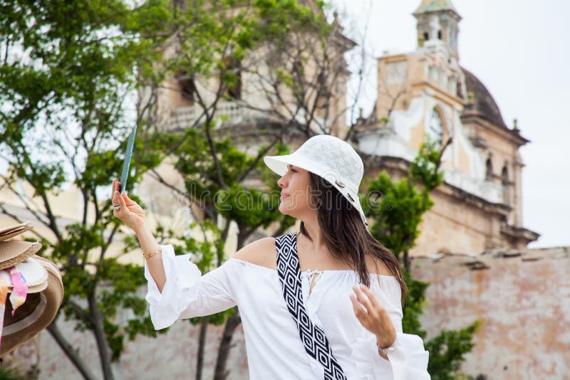 A jovem mulher bonita que tenta em chapéus comprar um de um vendedor ambulante em Cartagena de Índia murou a cidade fotografia de stock royalty free A jovem mulher bonita que tenta em chapéus comprar um de um vendedor ambulante em Cartagena de Índia murou a cidade fotografia de stock royalty free