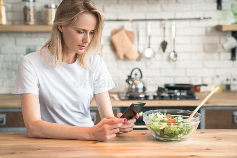 Jovem mulher bonita que usa o telefone celular ao fazer a salada na cozinha Alimento saud?vel Salada vegetal Dieta Estilo de vida foto de stock Jovem mulher bonita que usa o telefone celular ao fazer a salada na cozinha Alimento saud?vel Salada vegetal Dieta Estilo de vida foto de stock