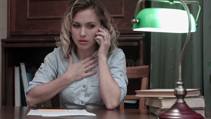 Jovem mulher cansado que senta-se em sua mesa que receiveing muito más notícias no telefone