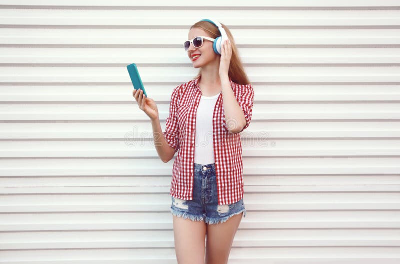 Jovem mulher de sorriso feliz nos fones de ouvido com smartphone que escuta a música que veste a camisa quadriculado, short no br imagem de stock royalty free Jovem mulher de sorriso feliz nos fones de ouvido com smartphone que escuta a música que veste a camisa quadriculado, short no br imagem de stock royalty free