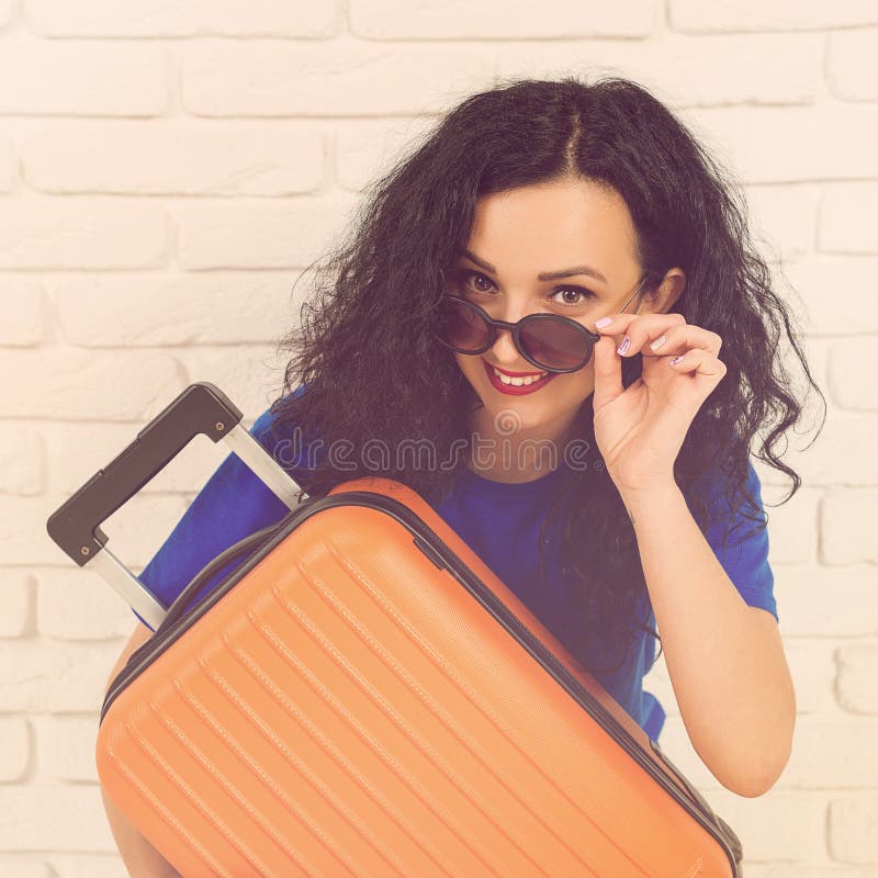 Jovem mulher de sorriso que abraça sua mala de viagem alaranjada A menina ocasional está pronta para viajar Mulher feliz nos ócul imagem de stock royalty free Jovem mulher de sorriso que abraça sua mala de viagem alaranjada A menina ocasional está pronta para viajar Mulher feliz nos ócul imagem de stock royalty free