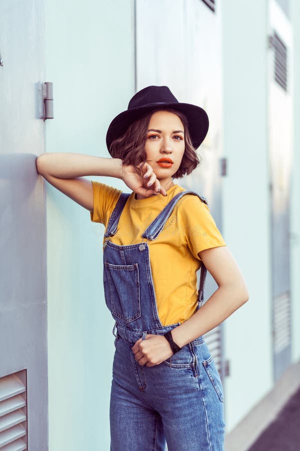 Jovem mulher em macac?es azuis da sarja de Nimes e no tshirt amarelo com a c?mera de vista sensual do chap?u negro ao levantar pe imagens de stock royalty free Jovem mulher em macac?es azuis da sarja de Nimes e no tshirt amarelo com a c?mera de vista sensual do chap?u negro ao levantar pe imagens de stock royalty free
