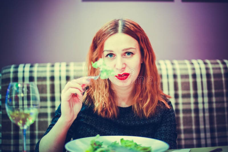 A jovem mulher está comendo salada vegetal misturada no café e bebe o vinho imagem de stock A jovem mulher está comendo salada vegetal misturada no café e bebe o vinho imagem de stock