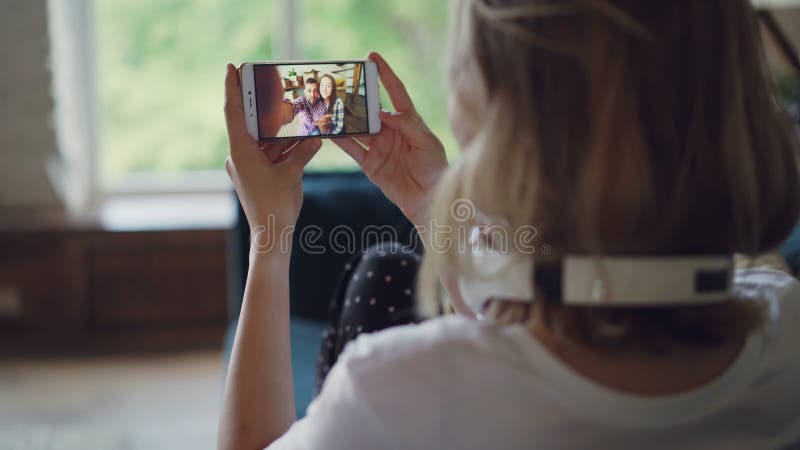 A jovem mulher está falando aos amigos em linha com o smartphone que olha a tela e que fala o assento no sofá em moderno