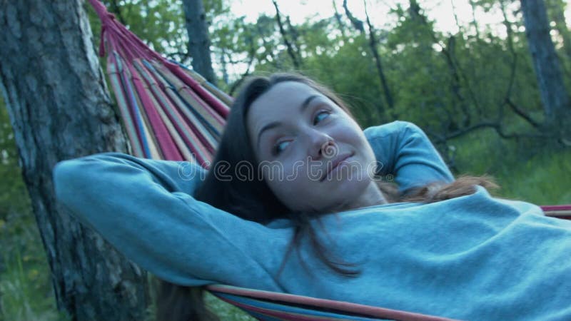 Jovem mulher feliz bonita em uma rede no sorriso do parque Na máscara das árvores, um balanço de descanso da menina na