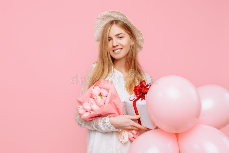 Jovem mulher feliz, em um vestido branco, estando com um ramalhete das flores, uma caixa de presente e balões, em um fundo cor-de fotografia de stock Jovem mulher feliz, em um vestido branco, estando com um ramalhete das flores, uma caixa de presente e balões, em um fundo cor-de fotografia de stock