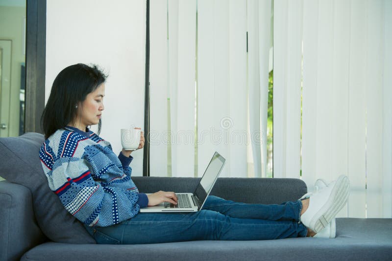 A jovem mulher feliz está relaxando no sofá confortável e está usando o portátil em casa Foto tonificada fotos de stock royalty free A jovem mulher feliz está relaxando no sofá confortável e está usando o portátil em casa Foto tonificada fotos de stock royalty free