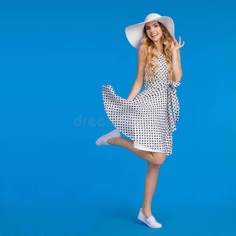 A jovem mulher feliz no vestido do verão, no chapéu de Sun e nas sapatilhas está estando em um pé fotografia de stock royalty free A jovem mulher feliz no vestido do verão, no chapéu de Sun e nas sapatilhas está estando em um pé fotografia de stock royalty free