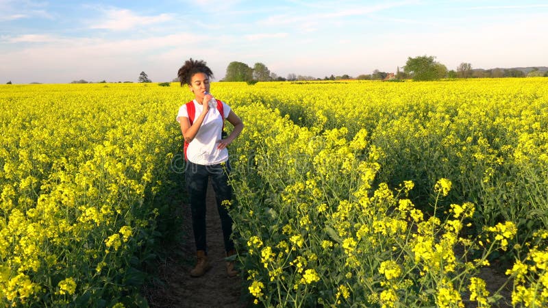 Jovem mulher fêmea do adolescente afro-americano da menina que caminha com trouxa e a garrafa vermelhas da água no campo da flor