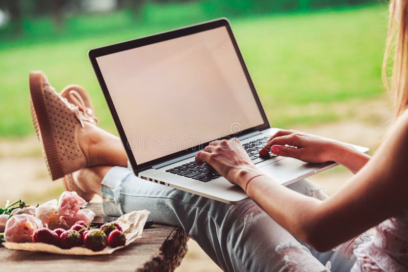 A jovem mulher laptop de utilização e de datilografia na tabela de madeira áspera com copo de café, morangos, ramalhete das peôni fotografia de stock A jovem mulher laptop de utilização e de datilografia na tabela de madeira áspera com copo de café, morangos, ramalhete das peôni fotografia de stock