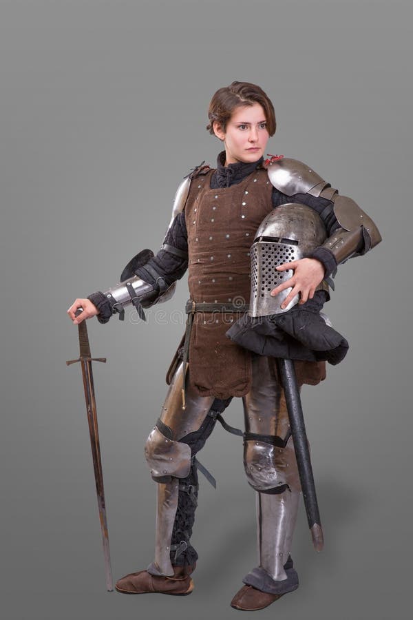 Jovem mulher no cavaleiro Armour que guarda o capacete em umas mão e espada em seu ombro sobre o fundo cinzento fotos de stock Jovem mulher no cavaleiro Armour que guarda o capacete em umas mão e espada em seu ombro sobre o fundo cinzento fotos de stock