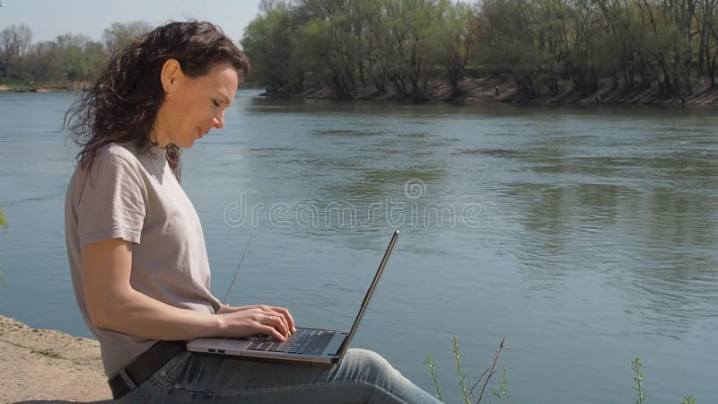 Jovem mulher no rio com um portátil A menina senta-se com um portátil no banco de rio