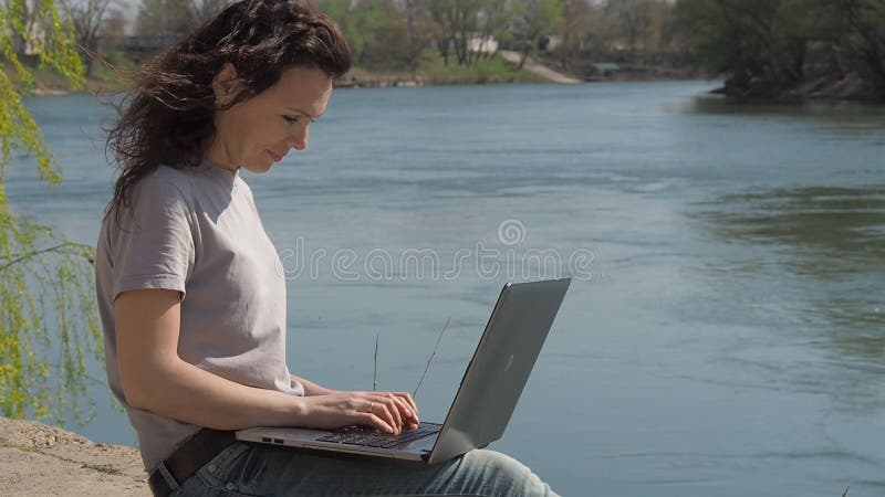 Jovem mulher no rio com um portátil A menina senta-se com um portátil no banco de rio