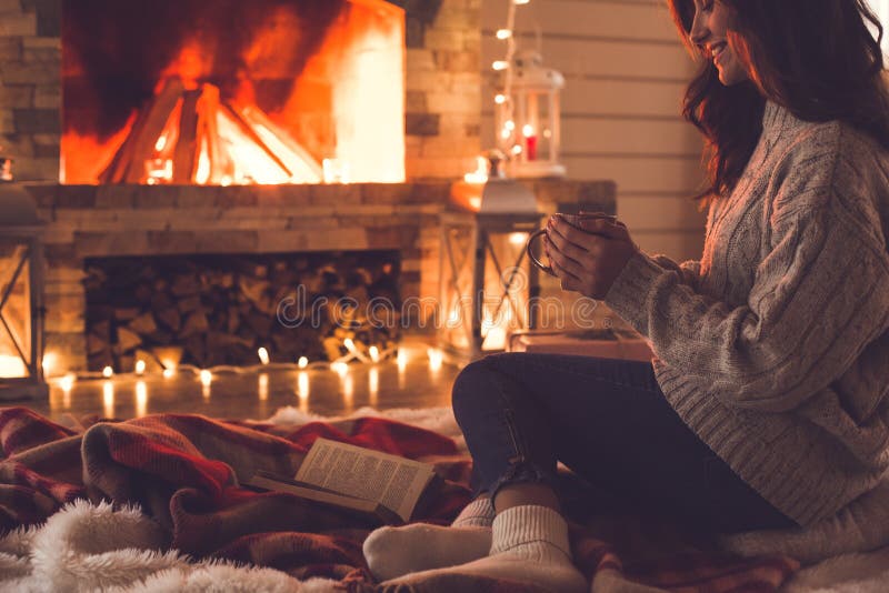 Jovem mulher perto do conceito do inverno da chaminé em casa que guarda o copo fotografia de stock Jovem mulher perto do conceito do inverno da chaminé em casa que guarda o copo fotografia de stock