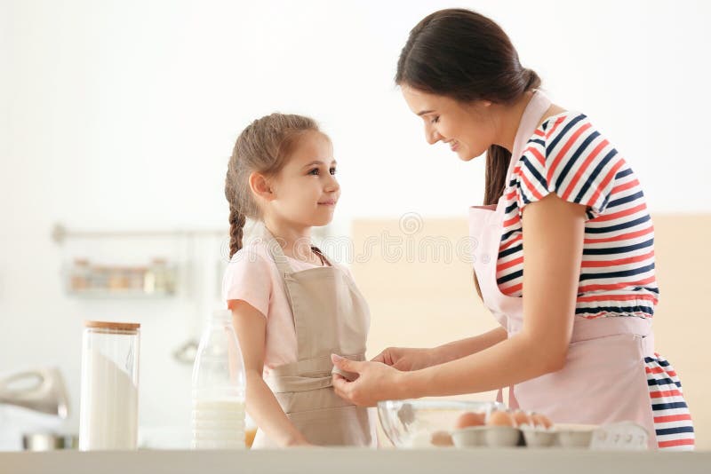 Jovem mulher que ajuda sua filha a pôr sobre o avental na cozinha Fazendo a massa junto fotografia de stock Jovem mulher que ajuda sua filha a pôr sobre o avental na cozinha Fazendo a massa junto fotografia de stock