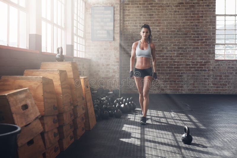 Full length shot of confident young woman walking in the crossfit gym. Muscular sportswoman warming up before a intense workout. Full length shot of confident young woman walking in the crossfit gym. Muscular sportswoman warming up before a intense workout.