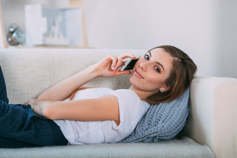 Jovem mulher que chama por seu telefone esperto que encontra-se no sofá foto de stock Jovem mulher que chama por seu telefone esperto que encontra-se no sofá foto de stock