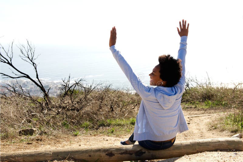 Rear view portrait of young woman sitting outdoors on a log with her hands raised in celebration. Rear view portrait of young woman sitting outdoors on a log with her hands raised in celebration