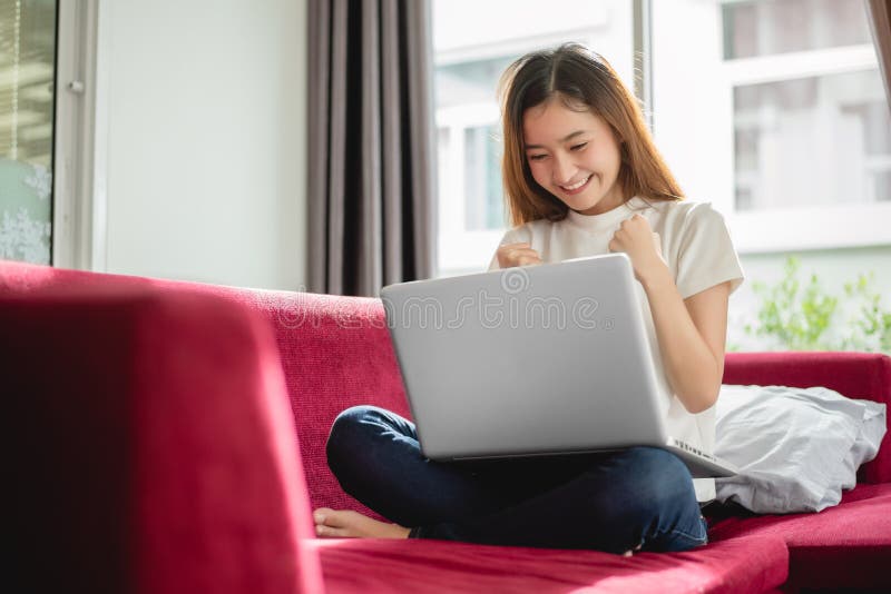 Jovem mulher que surfam o Internet pelo portátil no sofá vermelho e contente na troca bem sucedida em sua casa Venda e compra em