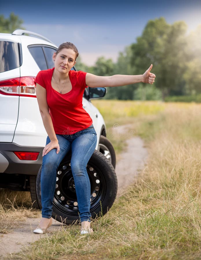 Jovem mulher virada com o carro quebrado que mantém o polegar ascendente e que viaja imagens de stock Jovem mulher virada com o carro quebrado que mantém o polegar ascendente e que viaja imagens de stock