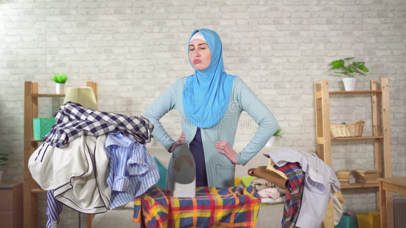 Jovem muçulmana cansada está engajada em passar uma pilha de roupa suja