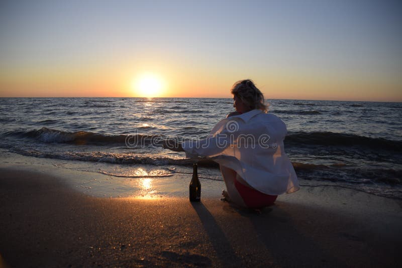 Jovem na praia marítima com uma garrafa de champanhe e um copo, mulher está sentada na areia na praia imagem de stock royalty free Jovem na praia marítima com uma garrafa de champanhe e um copo, mulher está sentada na areia na praia imagem de stock royalty free