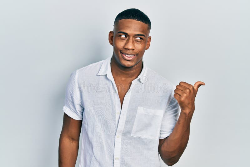 Young black man wearing casual white shirt smiling with happy face looking and pointing to the side with thumb up. Young black man wearing casual white shirt smiling with happy face looking and pointing to the side with thumb up