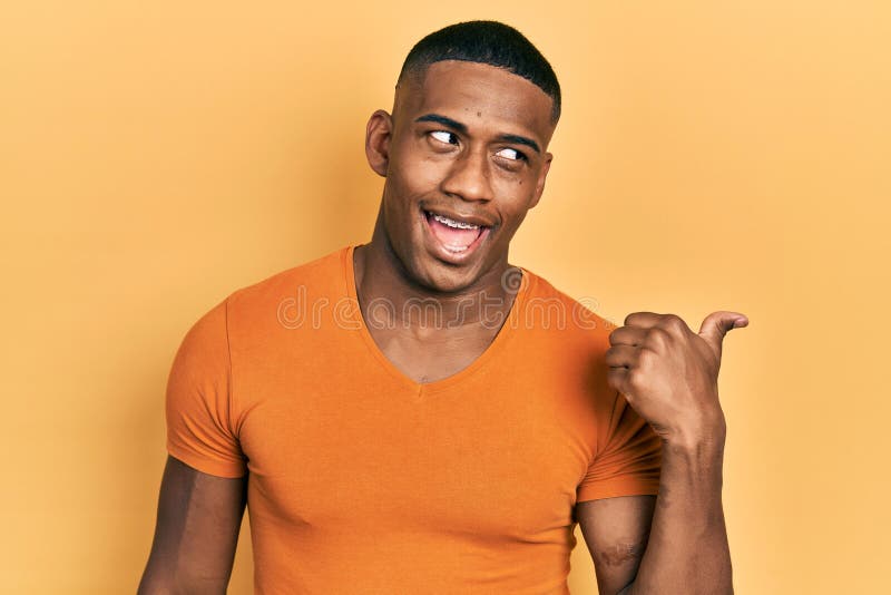 Young black man wearing casual orange t shirt smiling with happy face looking and pointing to the side with thumb up. Young black man wearing casual orange t shirt smiling with happy face looking and pointing to the side with thumb up