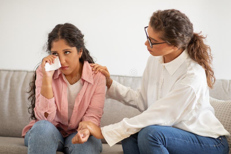 A young women appears visibly upset, wiping her tears with a tissue as she sits on a grey sofa. Beside her, a supportive psychologist is placing a comforting hand on her shoulder. A young women appears visibly upset, wiping her tears with a tissue as she sits on a grey sofa. Beside her, a supportive psychologist is placing a comforting hand on her shoulder