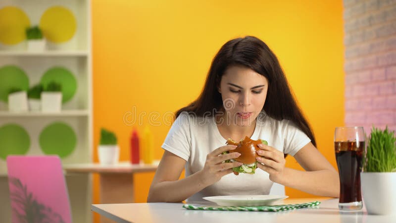 Jovem senhora com fome que come o cheeseburger saboroso no café, vidro do refresco na tabela