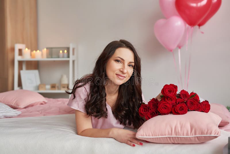 Jovem sexy na cama de pijama rosa com buquê de rosas vermelhas, quarto decorado com balões Cartão postal Feliz Dia dos Namorados, fotografia de stock Jovem sexy na cama de pijama rosa com buquê de rosas vermelhas, quarto decorado com balões Cartão postal Feliz Dia dos Namorados, fotografia de stock