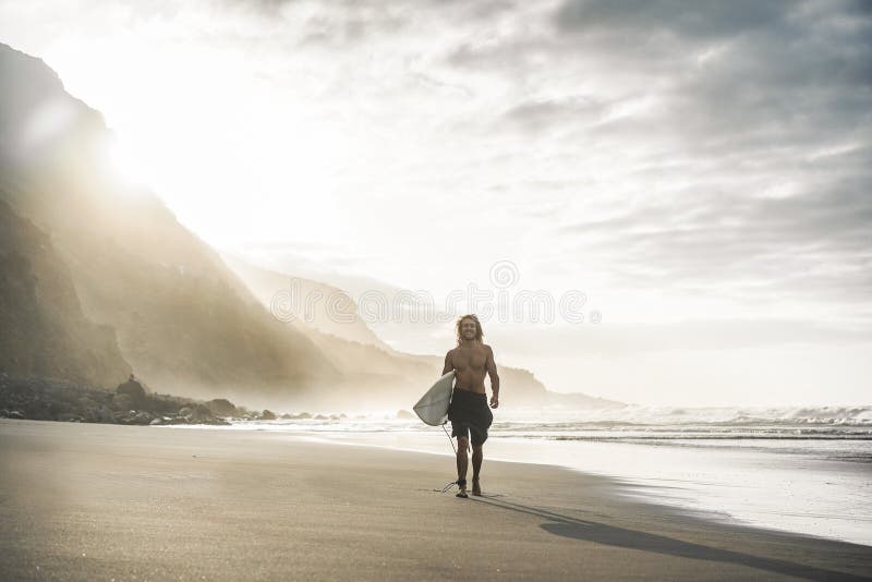 Young surfer on tropical beach at sunset - Man guy with his surf board walking - next the ocean on sunny day - Extreme sport - surfboard - man happy and laughing. Young surfer on tropical beach at sunset - Man guy with his surf board walking - next the ocean on sunny day - Extreme sport - surfboard - man happy and laughing