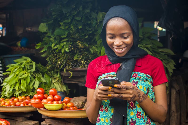 Joven africana vendiendo en un mercado local sonriendo mientras usa su teléfono móvil imagenes de archivo