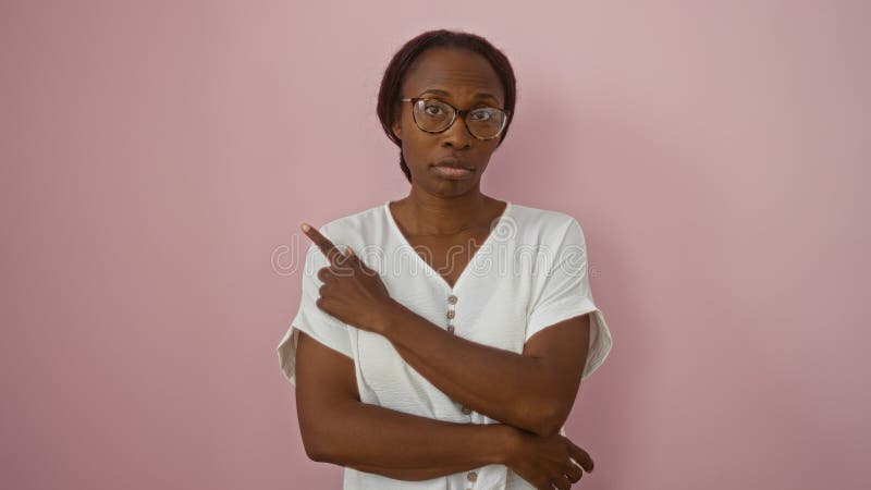 Joven afroamericana apuntando sobre un fondo rosado aislado con camisa blanca que se ve seria y considerada foto de archivo