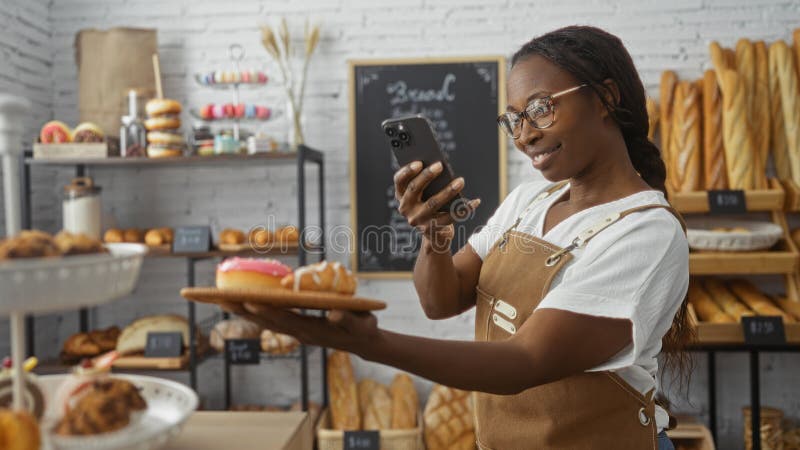 Joven afroamericana en una panadería tomando una foto de bollería con su teléfono rodeado de estantes de pan y dulces imagen de archivo libre de regalías