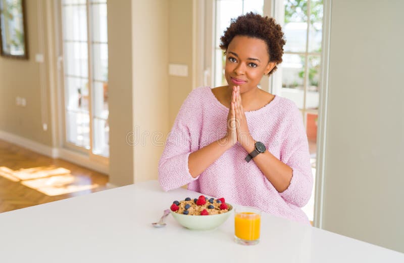 Joven afroamericana tomando un saludable desayuno en casa rezando con las manos juntas pidiendo perdón. Joven afroamericana tomando un desayuno saludable en casa rezando con las manos juntas pidiendo perdón sonriendo confiada