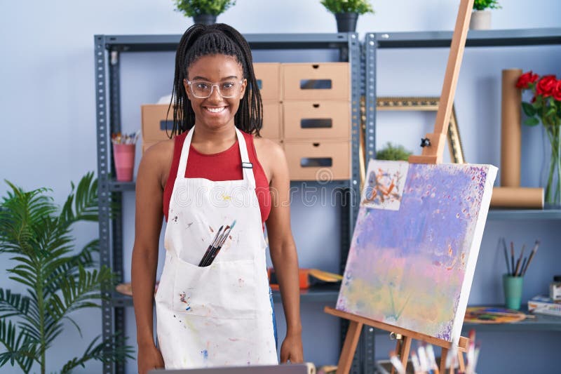 Joven afroamericano con trenzas en estudio de arte pintando lienzo con aspecto positivo y feliz y sonriendo con una fotos de archivo libres de regalías