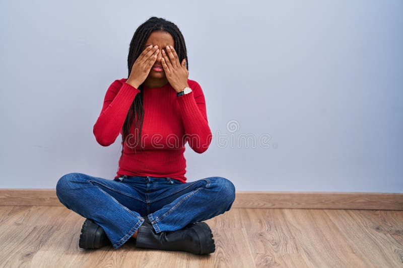 Joven afroamericano con trenzas sentadas en el suelo en casa frotando los ojos por fatiga y dolor de cabeza expresión somnolienta imagen de archivo