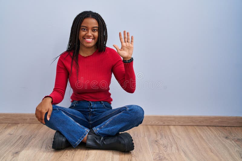 Joven afroamericano con trenzas sentadas en el suelo en su casa y apuntando con los dedos número cinco mientras sonríe fotografía de archivo libre de regalías
