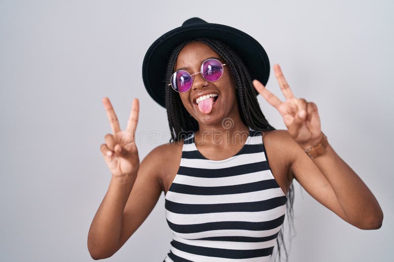 Joven afroamericano con trenzas con sombrero y gafas de sol sonriendo con la lengua mostrando los dedos de ambas manos haciendo la foto de archivo