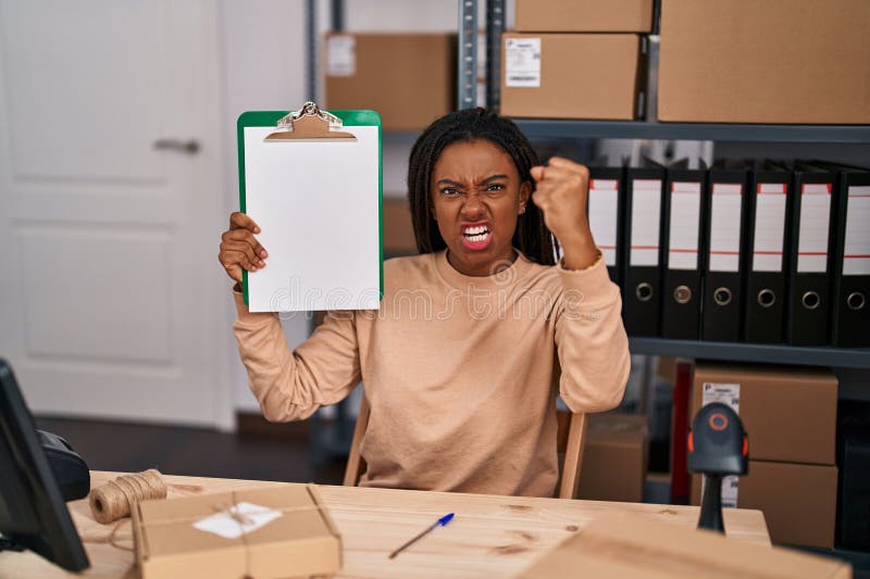 Joven afroamericano con trenzas trabajando en ecommerce de pequeñas empresas mostrando el portapapeles molesto y frustrado gritand imagen de archivo