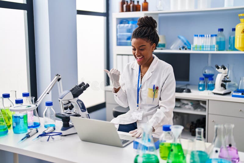 Joven afroamericano con trenzas trabajando en un laboratorio científico con una laptop apuntando al pulgar hacia el costado sonrie imágenes de archivo libres de regalías
